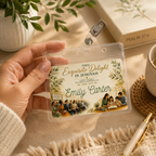 A hand holds a clear plastic ID badge holder with a printed personalized card that reads 'Find Exquisite Delight in Jehovah' and 'Emily Carter,' decorated with botanical illustrations and small group photos. The scene is styled on a beige fabric surface with a mug, pen, book, and plant in the background. Close-up of a personalized Circuit Assembly badge with premium clear holder and swivel clip.