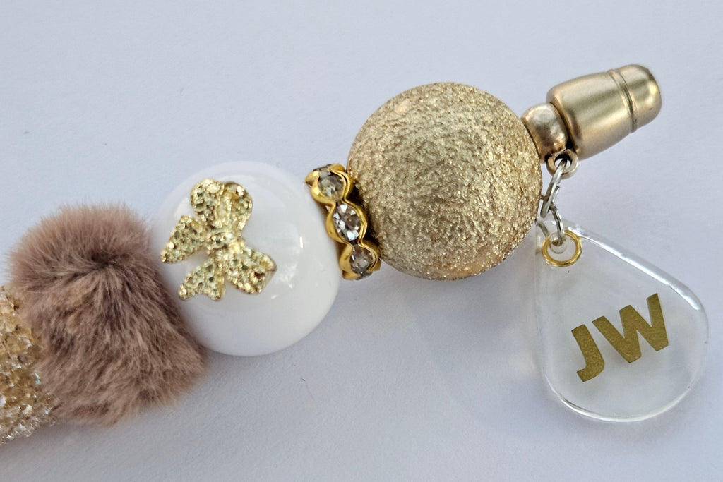 Decorative pen gold and white beads and a fluffy pom-pom on a light gray background. with JW charm