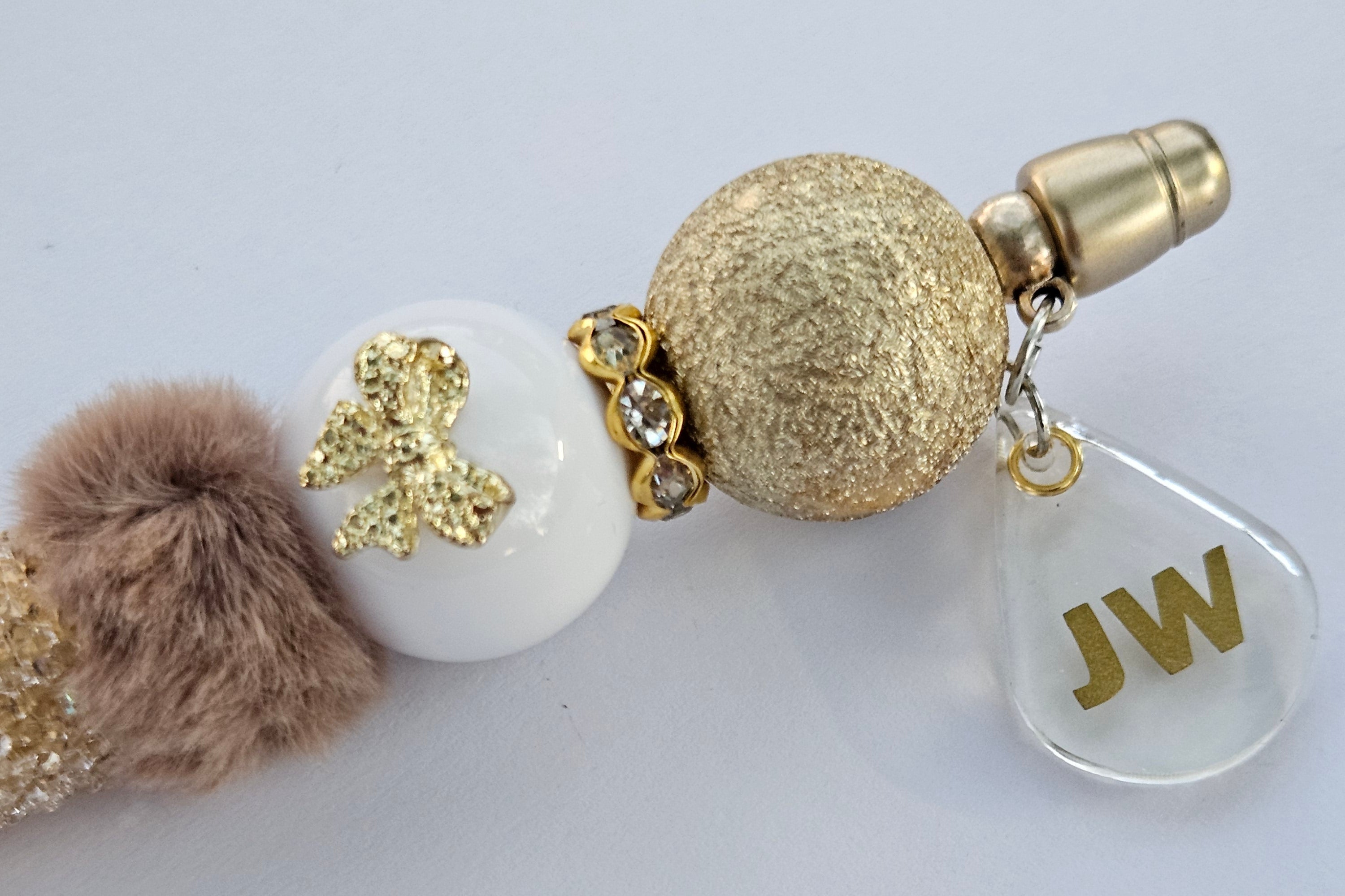 Decorative pen gold and white beads and a fluffy pom-pom on a light gray background. with JW charm