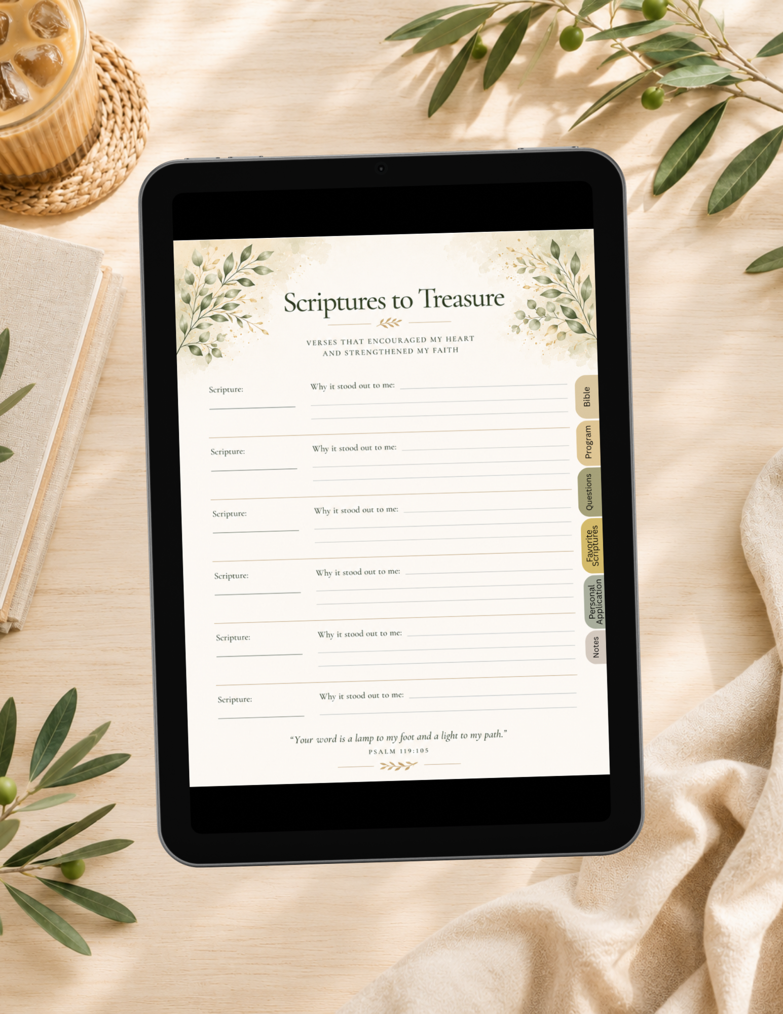 A tablet lying on a beige fabric surface, displaying a printable or digital planner page titled "Scriptures to Treasure" with lined writing areas and small decorative botanical borders. The scene is styled with olive leaves, a woven coaster, a notebook, and draped fabric.