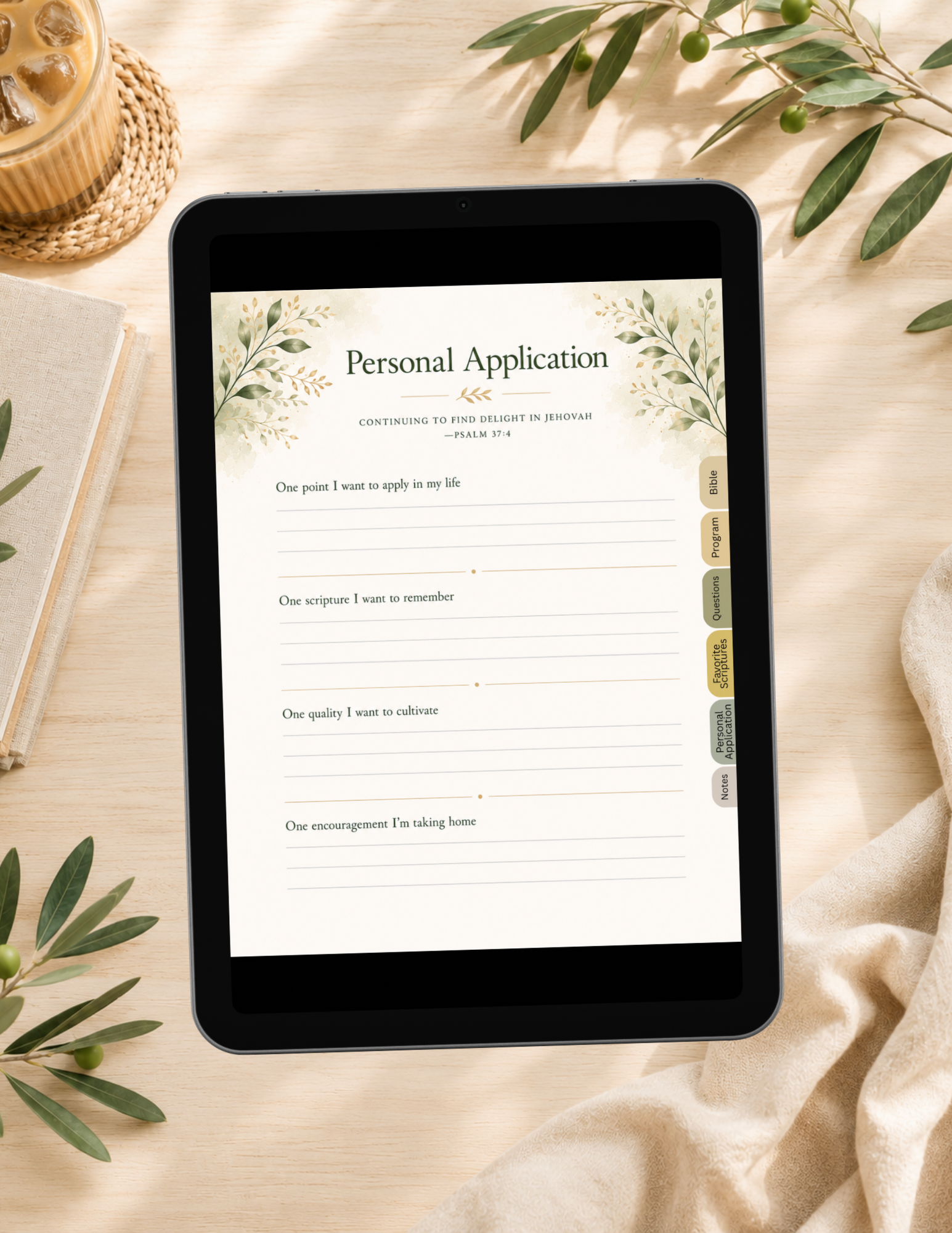 A tablet lying on a beige fabric surface, displaying a printable personal application form with floral watercolor accents. The scene is styled with a notebook, woven coaster, candle, and olive branches around it.