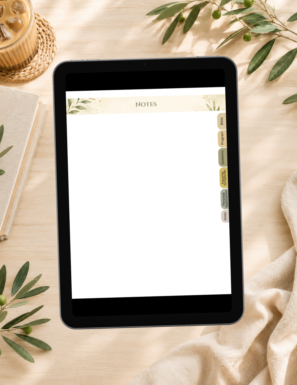 A tablet device displaying a notes app with a blank page and decorative header, placed on a light wood surface with a notebook, woven coaster, greenery, and draped fabric around it.