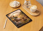 Tablet on a wooden surface with a scenic cover, next to a glass of water and a cup of coffee.