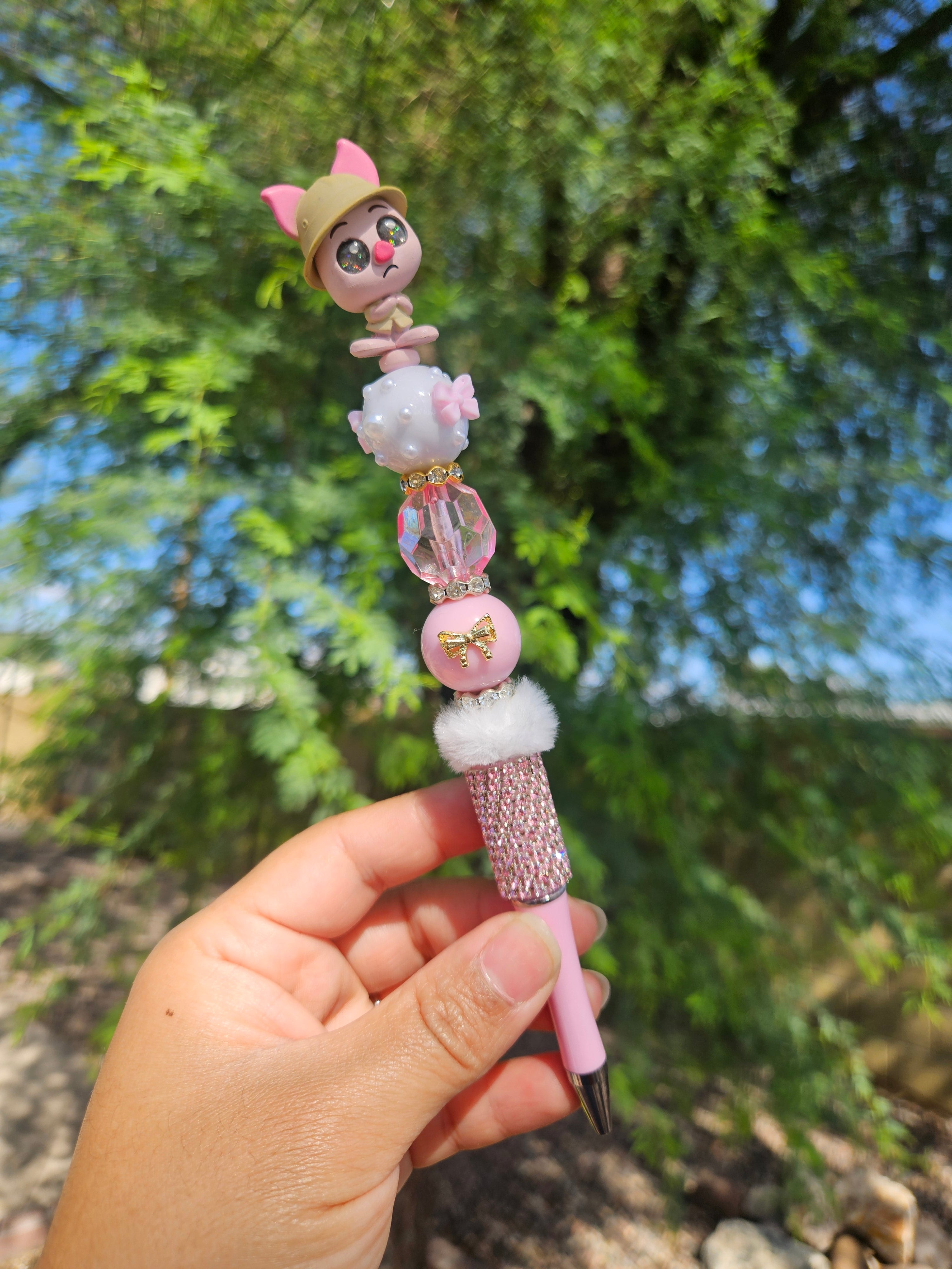 Hand holding a piggy beadable pen with a blurred green outdoor background